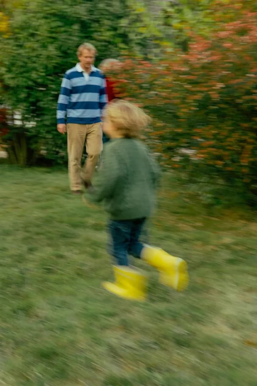 A child in yellow rain boots running through a garden toward grandparents, in motion.
